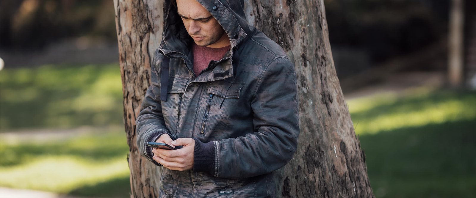 A young man wearing a dark hooded jacket, standing against a tree texting on a phone.