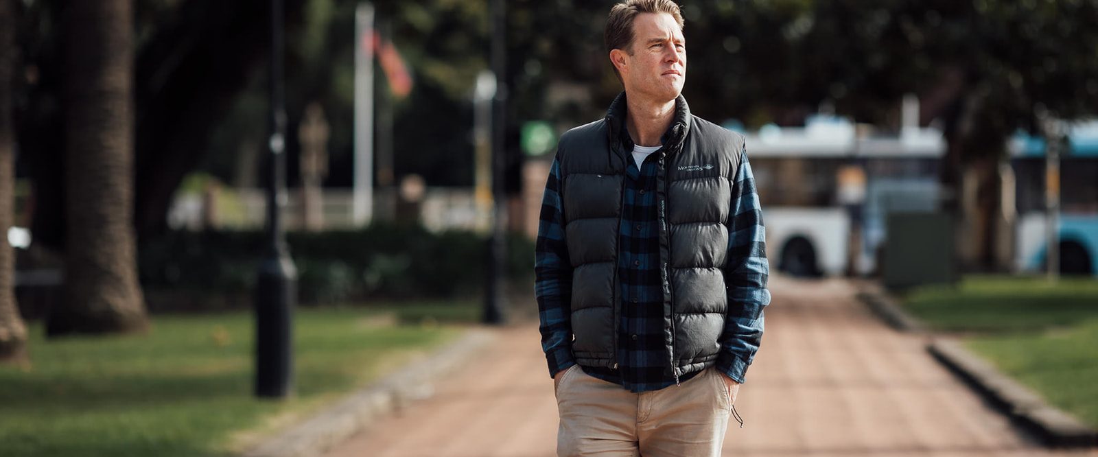 A troubled looking man wearing a checked shirt and black puffy vest walking in a park.