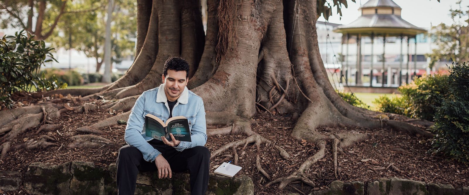 A man smiling and reading a book, sitting under a tree.